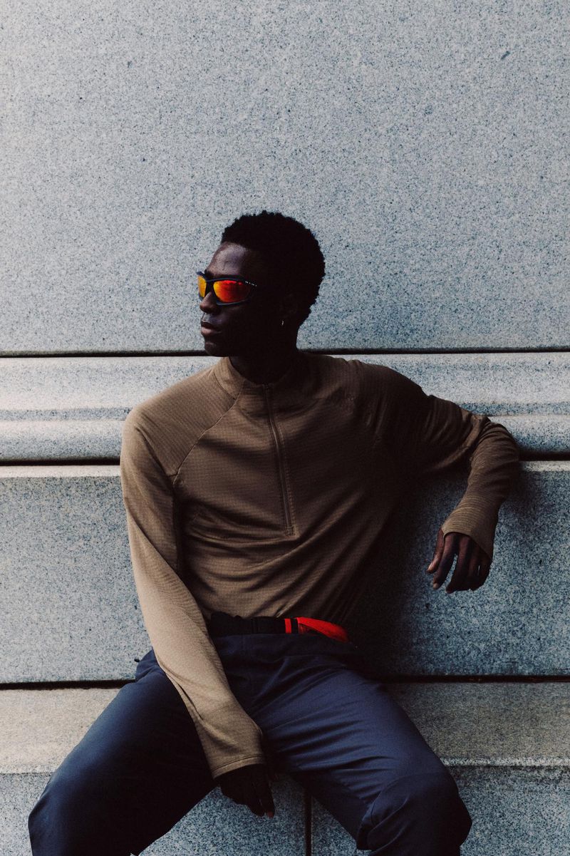 A Black man wearing sunglasses and athletic wear sits on a concrete wall, angled to the left in an outdoor setting with natural light.