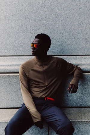 Feranmi Ayeni in a brown half-zip long sleeve top, dark trousers, and mirrored orange sunglasses leaning against a stone wall.