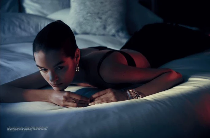 A woman with dark hair and jewelry poses while lying on a bed with white linens, showcasing fashion and beauty in a dimly lit indoor setting.