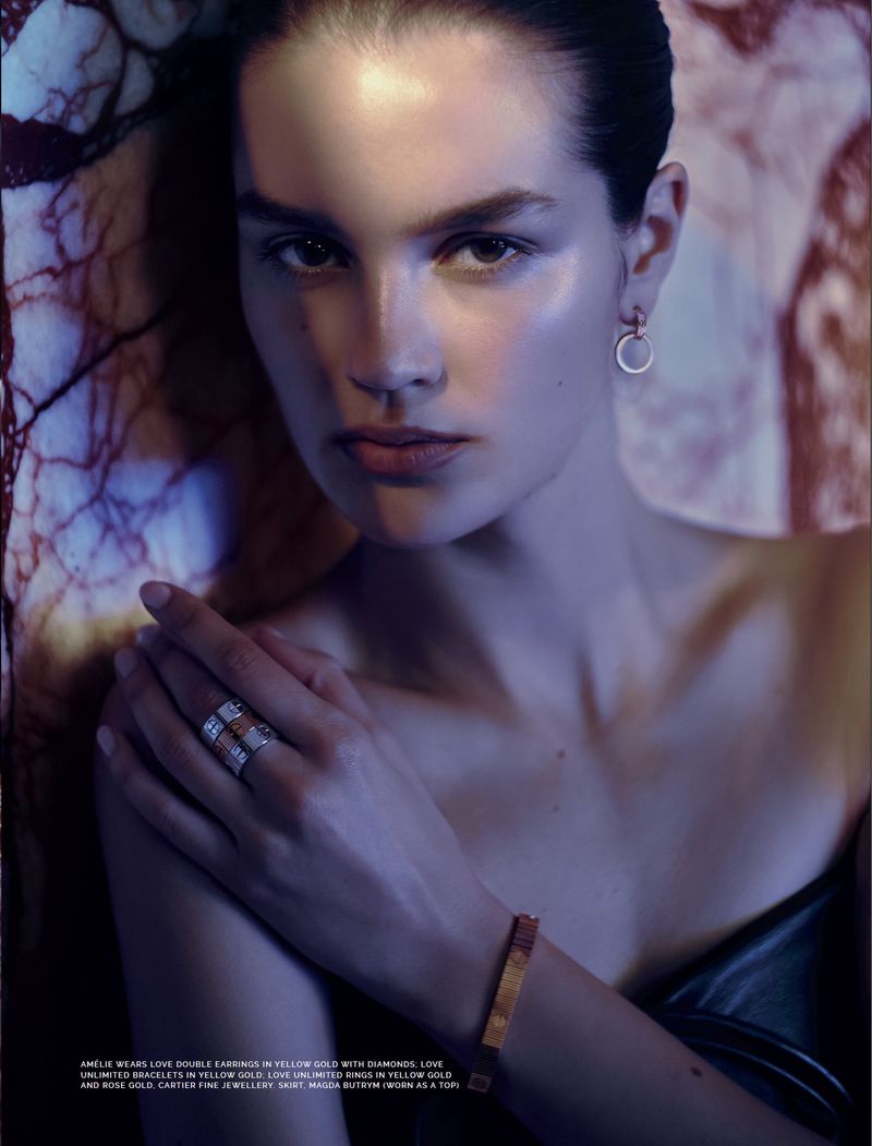 Close-up studio portrait of a woman with dark hair wearing jewellery including hoop earrings, rings, and bracelet, against a mottled background.