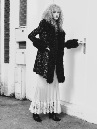 Monochrome fashion portrait of a woman with curly hair wearing a dark textured coat and white lace skirt beside an exterior door.