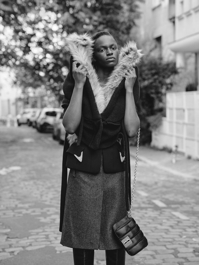 Black and white portrait of an adult woman wearing a fur collared coat with a skirt and holding a purse on a cobblestone street.