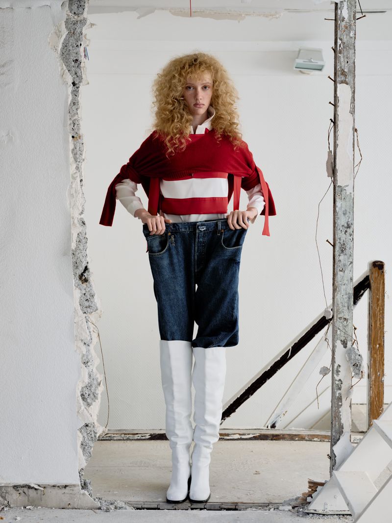 Fashion shot of a woman with curly hair wearing a red top and white boots, posing in a doorway of a destroyed white wall indoors.