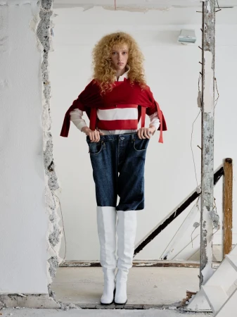 Fashion shot of a woman with curly hair wearing a red top and white boots, posing in a doorway of a destroyed white wall indoors.