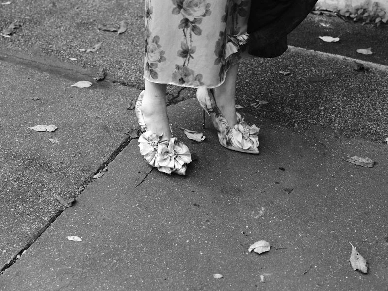 Close-up of woman's legs wearing floral dress and floral-embellished heels, standing on a wet urban street with fallen leaves.