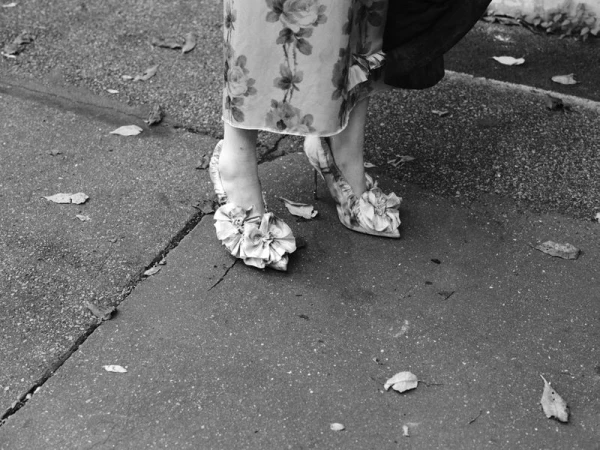 Close-up of woman's legs wearing floral dress and floral-embellished heels, standing on a wet urban street with fallen leaves.