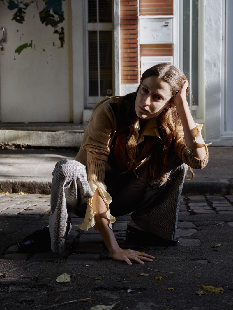 A young woman with long brown hair poses while crouching on a cobblestone street next to a building with a white door and brick wall.