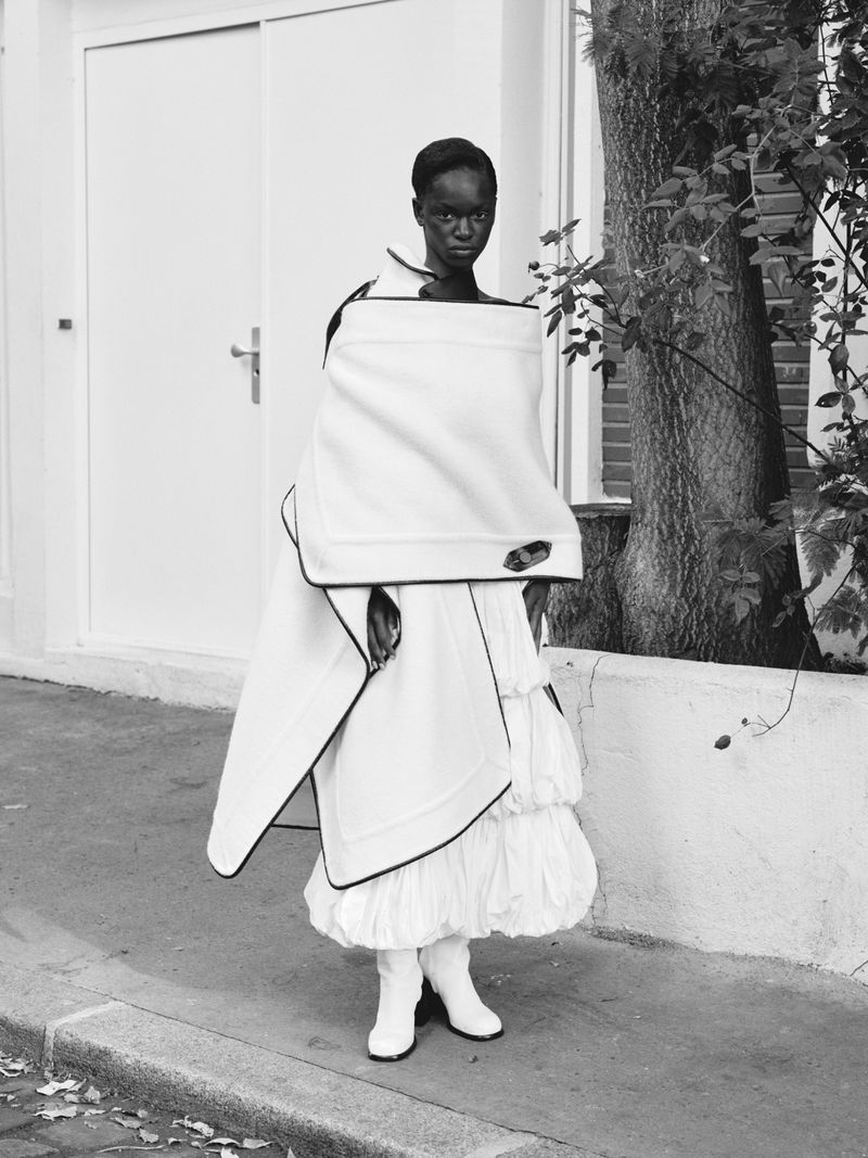Fashion shot of an African woman in a white outfit with black trim, posing outdoors next to a white wall and a tree.