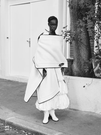 Fashion shot of an African woman in a white outfit with black trim, posing outdoors next to a white wall and a tree.
