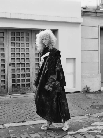 Model with voluminous crimped blonde hair wearing an oversized dark draped coat and pointed high heels standing on a paved street.