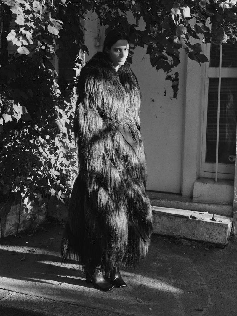 A woman poses in a long, textured, dark coat, standing outside a building with greenery in a black-and-white fashion photograph.