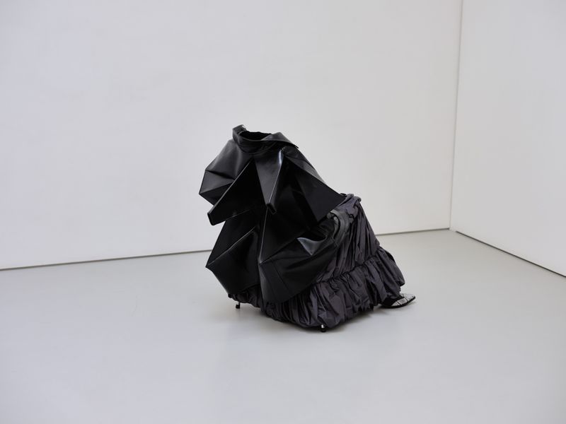 Abstract black dress sculpture in a white corner room, featuring shiny textures and dramatic folds, creating a unique display.
