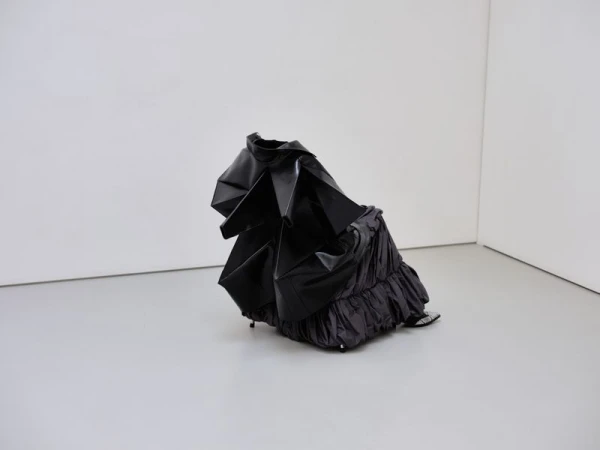 Abstract black dress sculpture in a white corner room, featuring shiny textures and dramatic folds, creating a unique display.