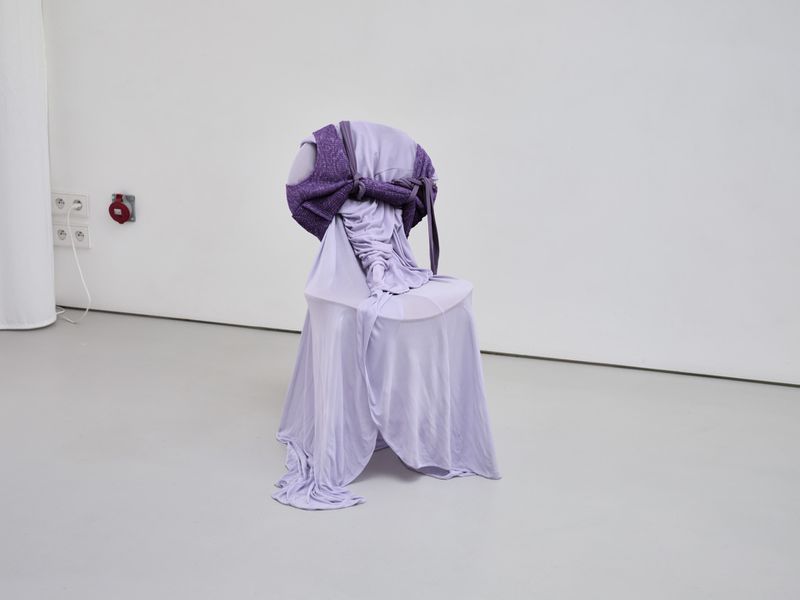 Chair draped in lilac fabric with a purple textured wrap, placed indoors against a white wall, creating a minimalist art installation.