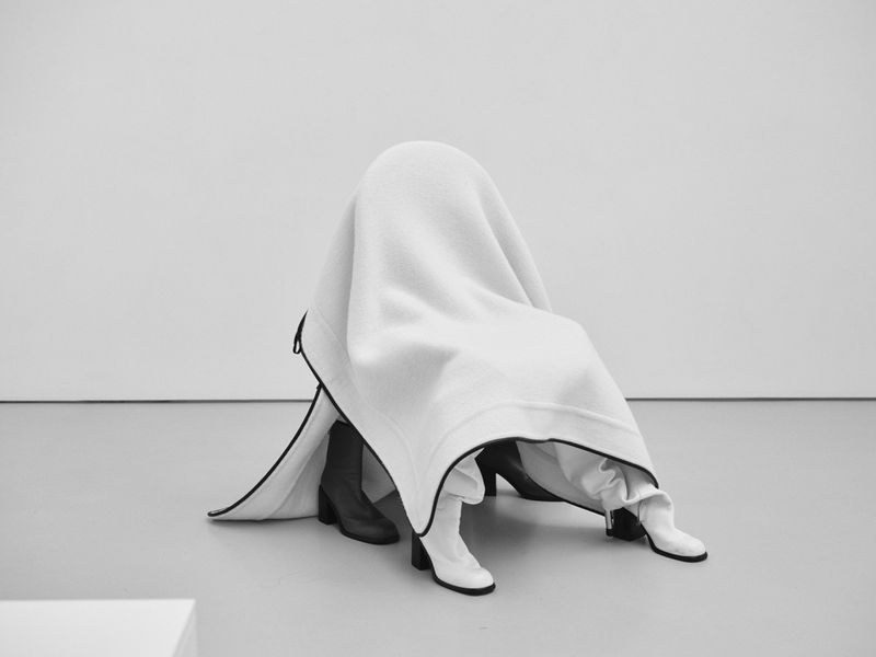 Person under white blanket wearing boots indoors against white background in studio shot. Contemporary style, minimalist.