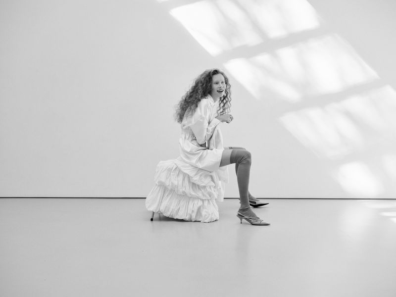 Black and white portrait of an adult female with curly hair wearing a white shirt and thigh-high socks sitting on a ruffled stool.