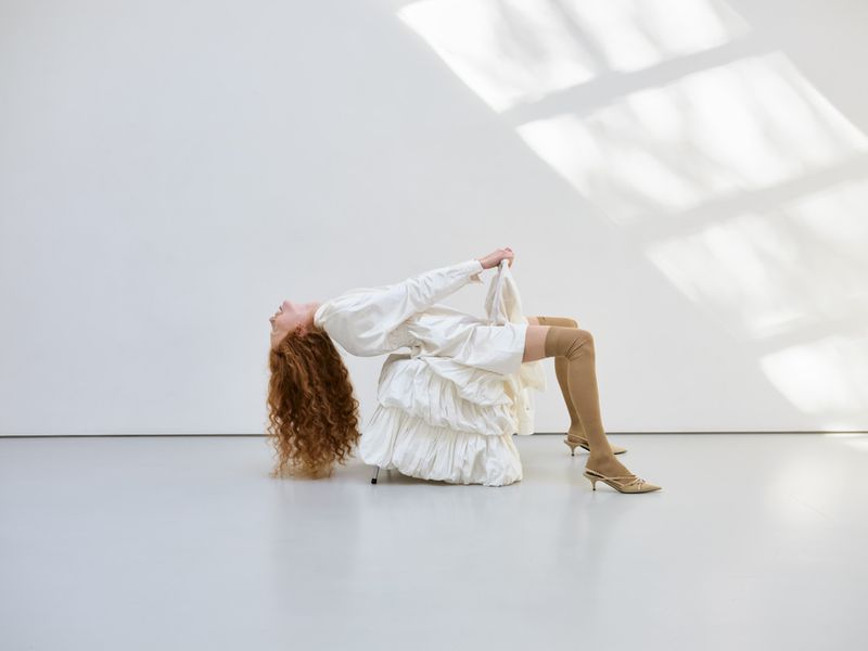 Woman with red hair in arched pose wearing white dress and tan thigh-high boots sitting on chair in front of bright white wall.