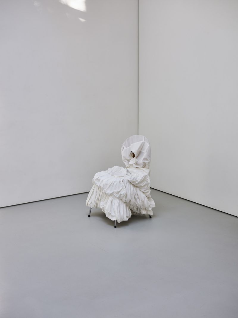 Unique ruffled chair on metal legs, placed against a minimal white wall and gray floor in a minimalist interior space.