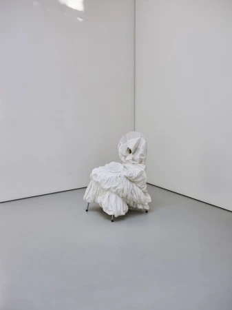 Unique ruffled chair on metal legs, placed against a minimal white wall and gray floor in a minimalist interior space.