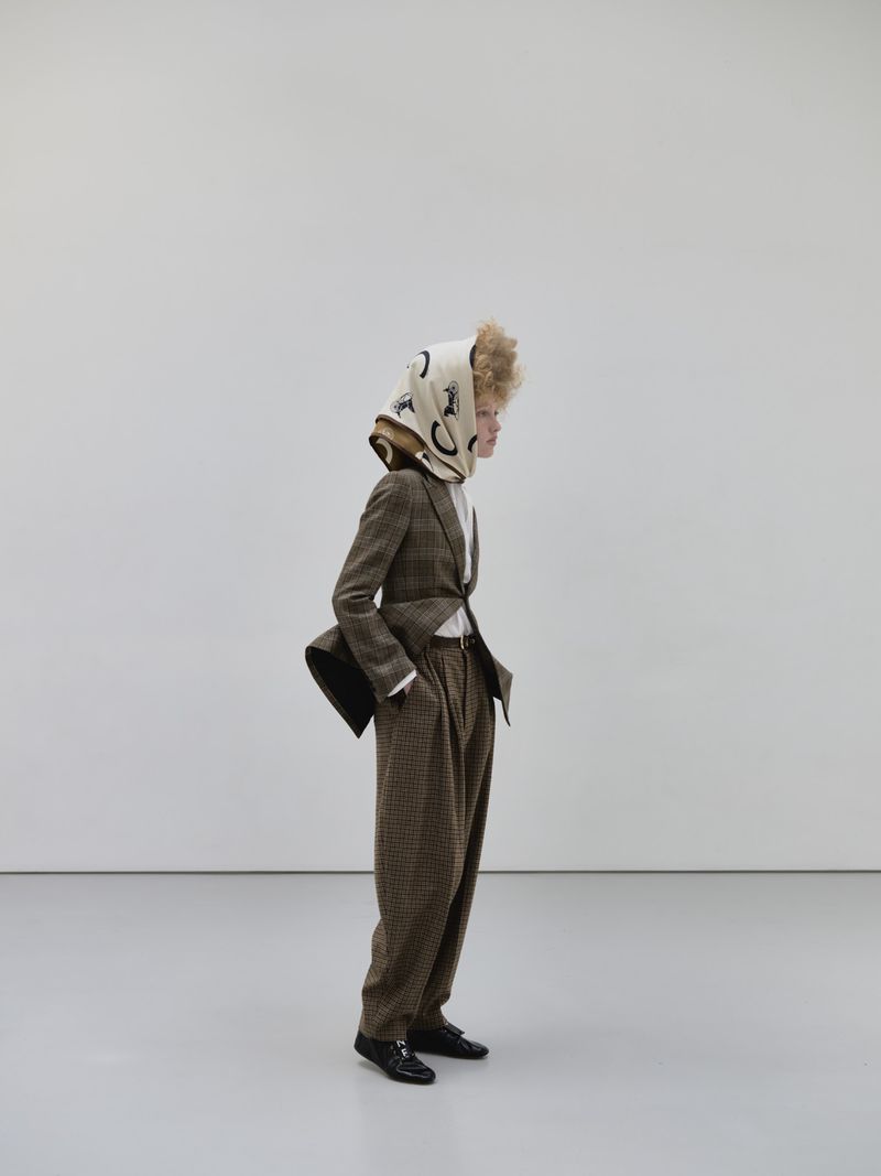 Model in a tailored brown suit and printed headscarf standing in profile against a neutral minimalist studio wall.