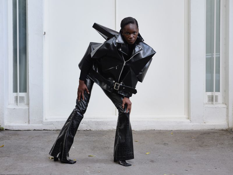 Fashion shot of a Black woman in a dramatic black leather jacket and flared pants posing in front of a white building, eye-level view.