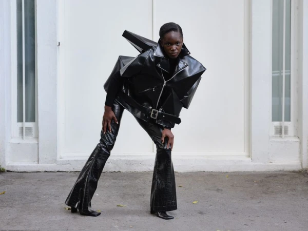 Fashion shot of a Black woman in a dramatic black leather jacket and flared pants posing in front of a white building, eye-level view.