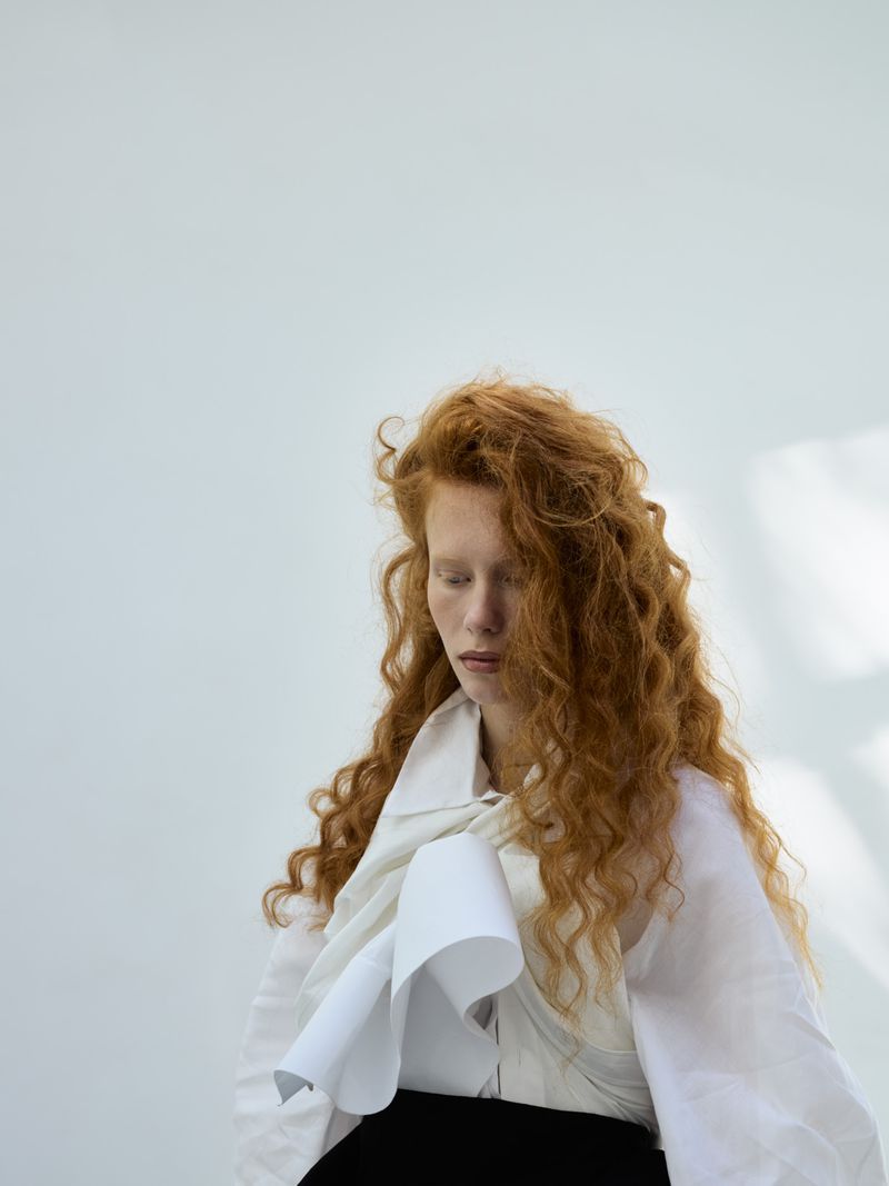 Portrait of a redhead woman with voluminous curls wearing a white top with a large collar and a black skirt, against a plain background.