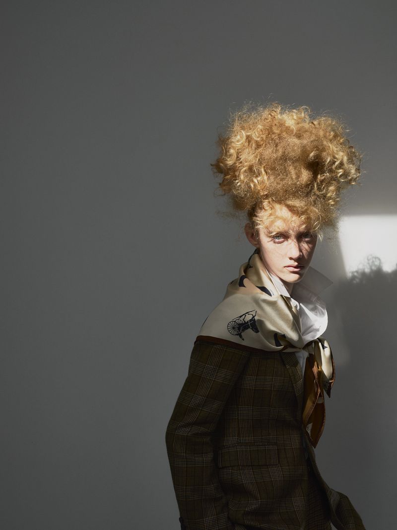 A young adult with a striking blonde afro poses in a plaid jacket and patterned scarf against a neutral gray background, illuminated by sunlight.