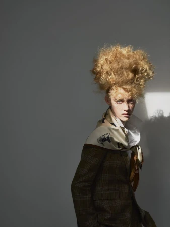 A young adult with a striking blonde afro poses in a plaid jacket and patterned scarf against a neutral gray background, illuminated by sunlight.