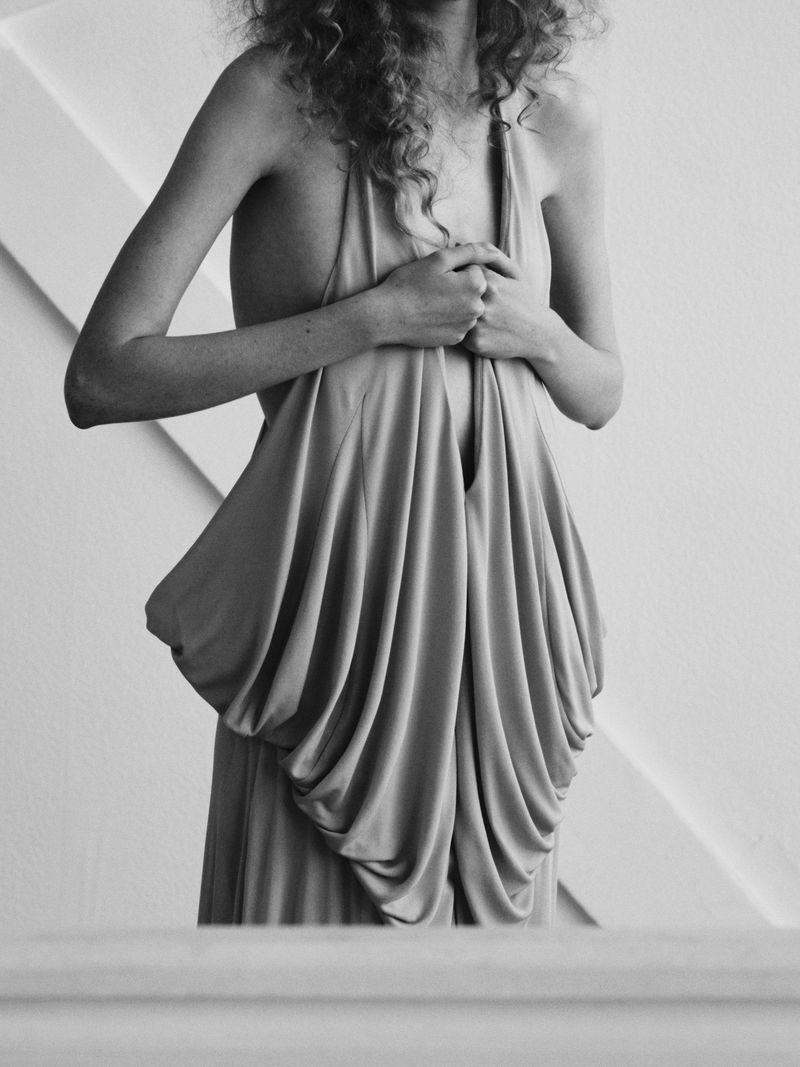 Black and white photo of an adult woman with curly hair wearing a draping dress, hands clasped in front of her, indoor setting.