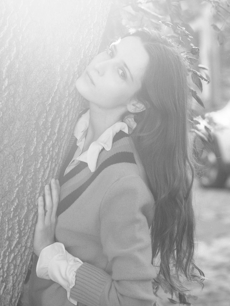Black-and-white portrait of a long-haired young woman in V-neck top posing next to a tree, with soft lighting and a natural outdoor setting.
