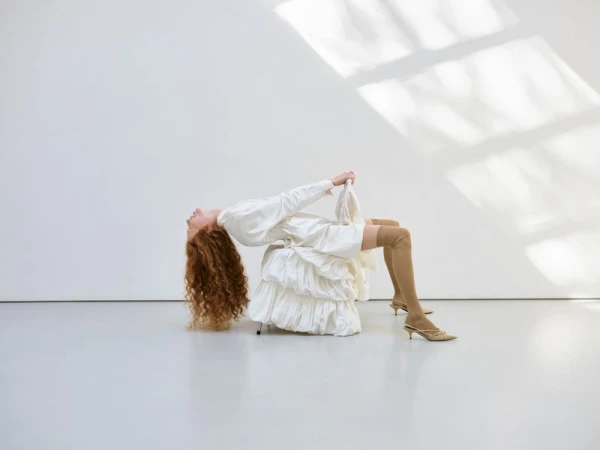 Woman with long red hair arching backward over a white ruffled seat wearing beige thigh-high socks and pointed kitten heels in a studio.