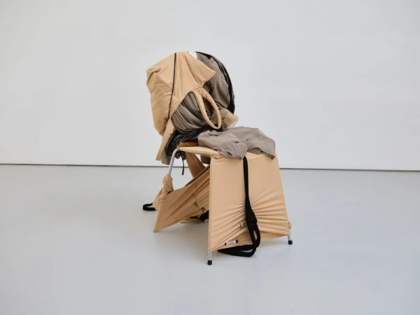 Sculptural modern chair wrapped in draped beige and taupe fabric garments on a light studio floor.