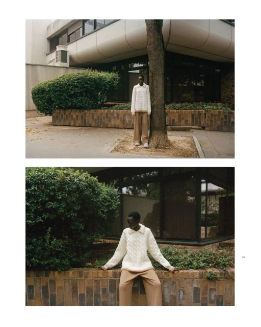 Fashion shot of a Black model wearing a cream-colored cable knit sweater and tan pants, posing outdoors next to a tree in an urban setting.