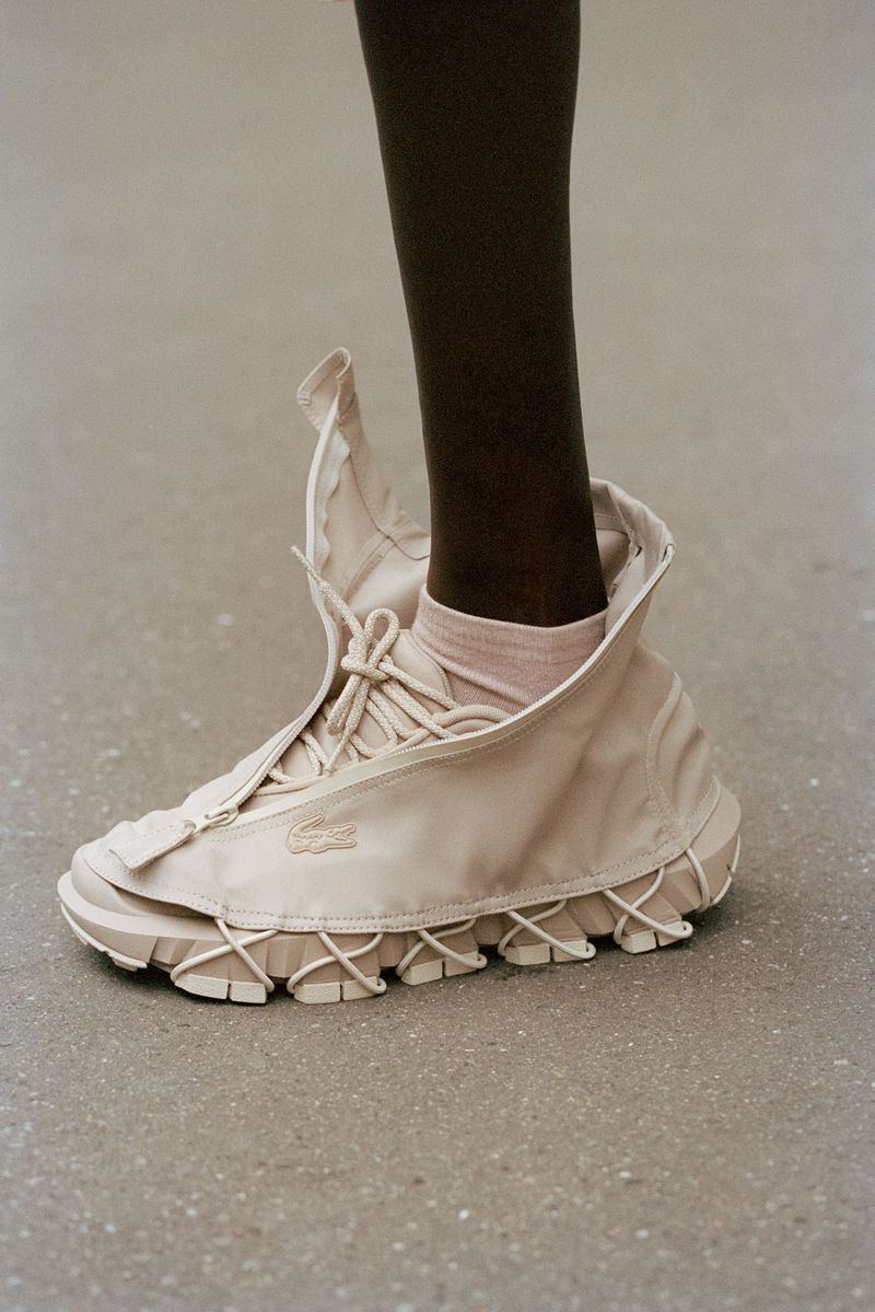 Tan Lacoste sneaker with a zipper opening shown on a person's leg standing on a light grey surface. The shoe is a high-top design.