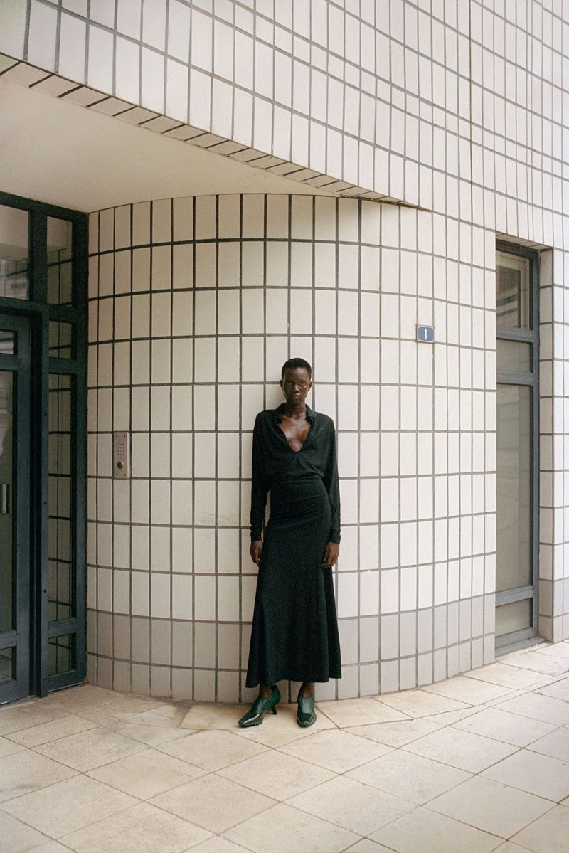 Woman poses against a tiled building in a wrap dress and heels. The architecture has a rounded corner and natural light.
