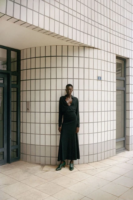Woman poses against a tiled building in a wrap dress and heels. The architecture has a rounded corner and natural light.