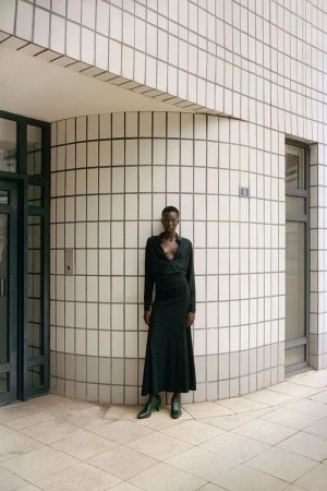 Woman poses against a tiled building in a wrap dress and heels. The architecture has a rounded corner and natural light.