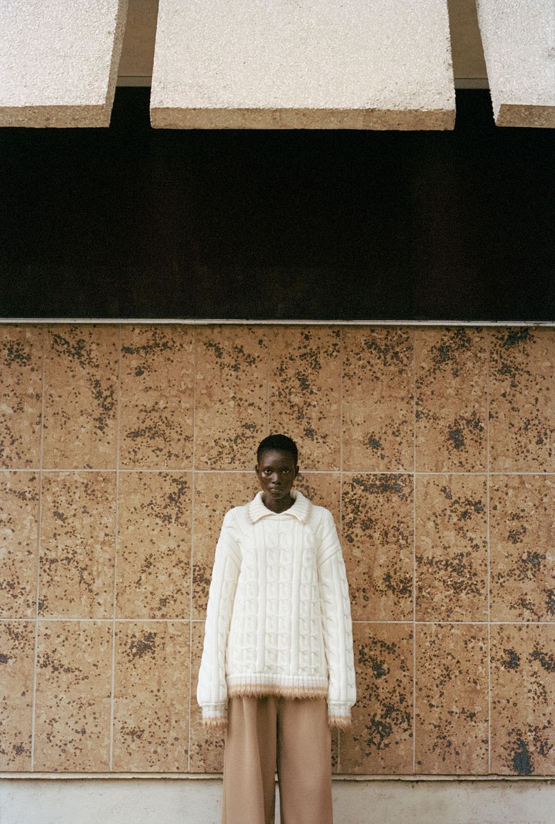 Adult female poses outdoors wearing a white, cable-knit sweater and wide-leg pants in front of a tiled wall and overhanging architecture.