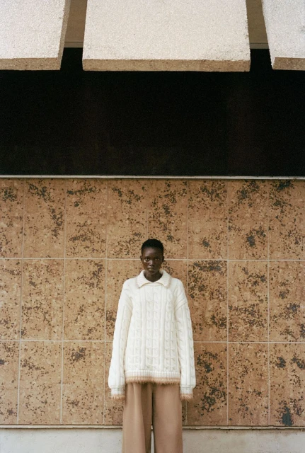 Adult female poses outdoors wearing a white, cable-knit sweater and wide-leg pants in front of a tiled wall and overhanging architecture.