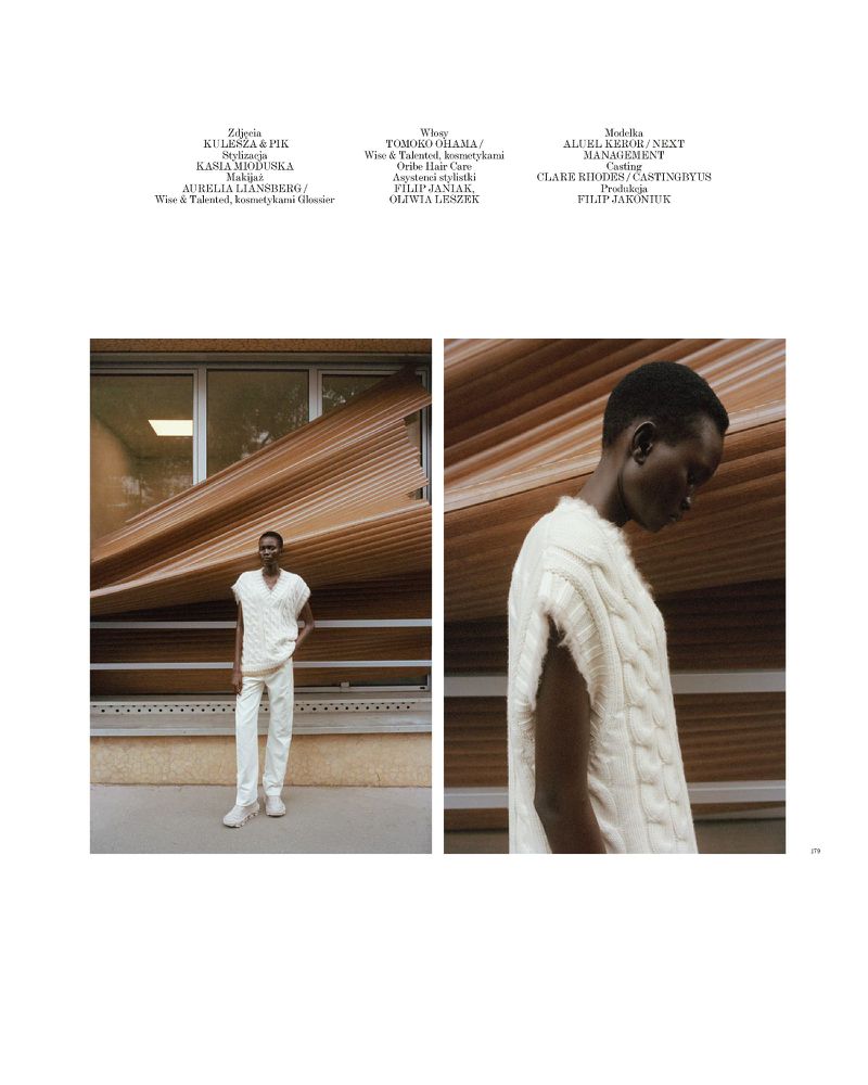 Fashion editorial shot of a dark-skinned model wearing knitted vest and pants, posing against a textured building facade in daylight.