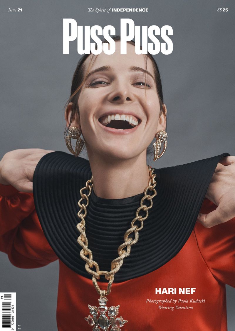 Magazine cover featuring Hari Nef with a wide smile, wearing a red top and oversized gold jewelry against a gray background.