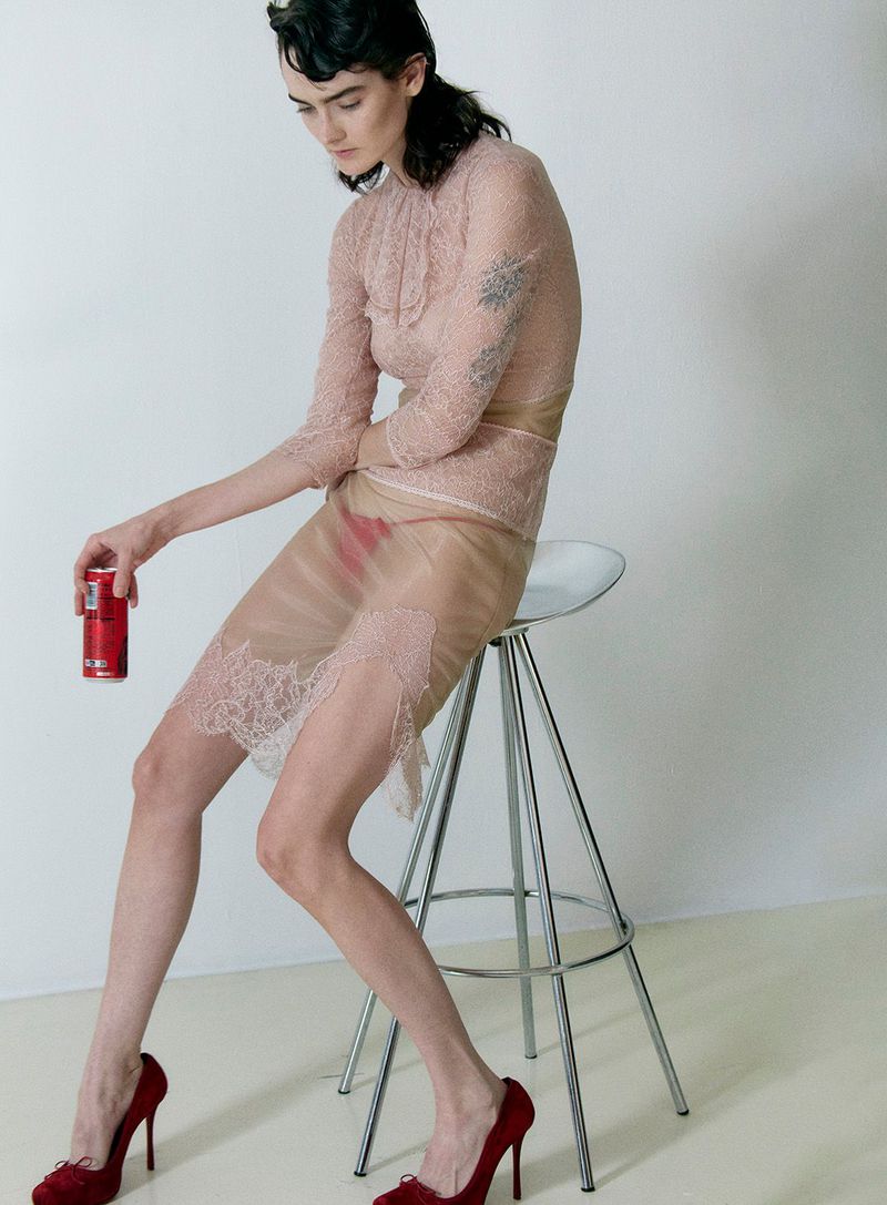An adult female sits on a clear stool in a studio holding a can of soda. She is wearing a sheer lace dress and red heels.