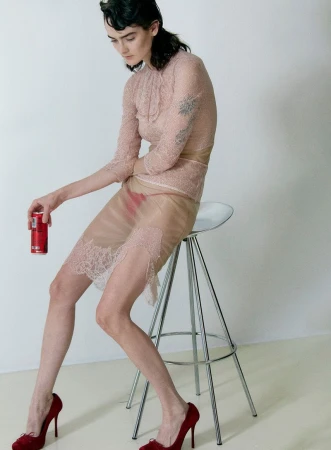 An adult female sits on a clear stool in a studio holding a can of soda. She is wearing a sheer lace dress and red heels.