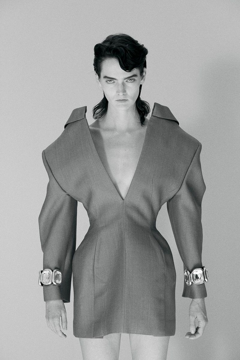 Fashion portrait of a woman wearing a V-neck grey dress with wide shoulders and chunky silver bracelets standing in a grey photo studio.