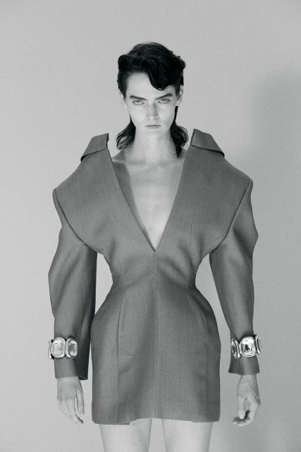Fashion portrait of a woman wearing a V-neck grey dress with wide shoulders and chunky silver bracelets standing in a grey photo studio.