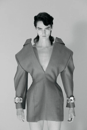 Fashion portrait of a woman wearing a V-neck grey dress with wide shoulders and chunky silver bracelets standing in a grey photo studio.