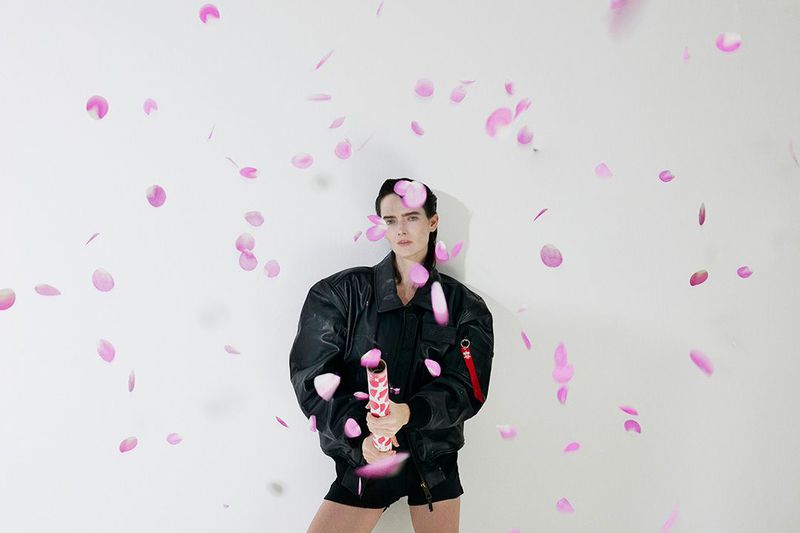 Woman in a black jacket poses with a party popper amidst falling pink petals against a white backdrop in a brightly lit photo studio.