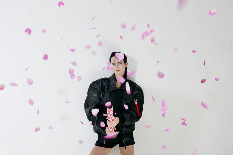 Woman in a black jacket poses with a party popper amidst falling pink petals against a white backdrop in a brightly lit photo studio.