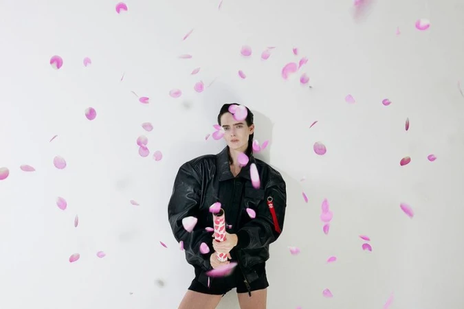 Woman in a black jacket poses with a party popper amidst falling pink petals against a white backdrop in a brightly lit photo studio.